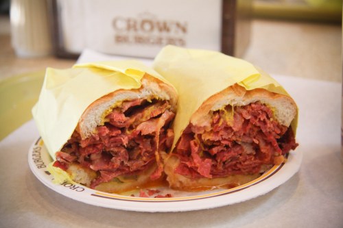 A close-up of a sandwich filled with layers of meat, wrapped in yellow paper, served on a plate.