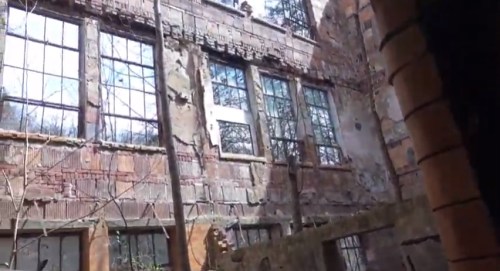 Abandoned building with crumbling walls and large, broken windows, surrounded by overgrown vegetation.