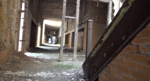 An abandoned building interior with exposed brick walls, wooden beams, and a staircase leading into a dimly lit corridor.