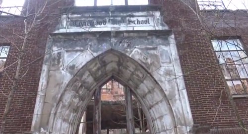 Entrance of an abandoned school building with a gothic arch and weathered brick walls, surrounded by overgrown vegetation.