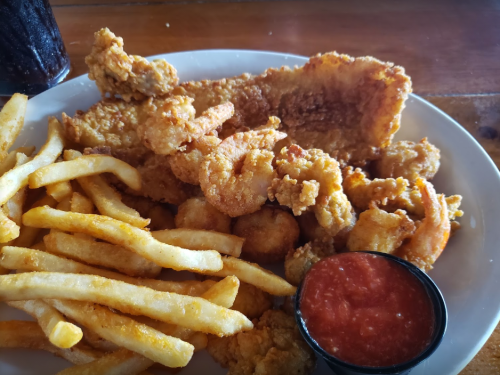A plate of fried seafood including shrimp, fish, and hushpuppies, served with fries and a side of dipping sauce.