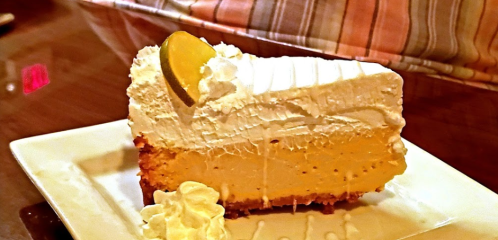 A slice of creamy cheesecake topped with whipped cream and a lemon wedge, served on a white plate.