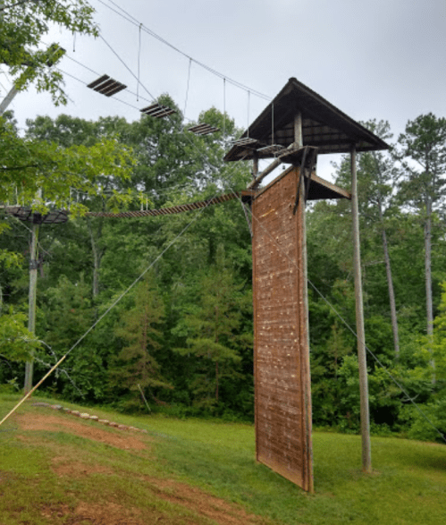 A tall wooden climbing wall with a rope course above, surrounded by trees and grass in a natural setting.