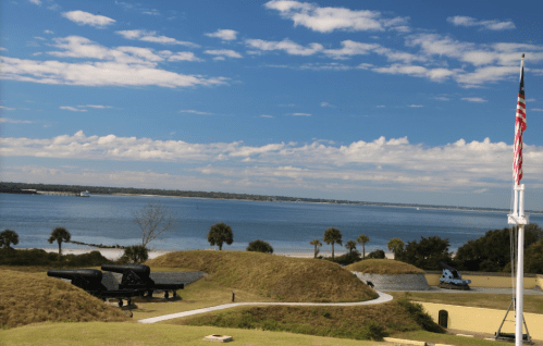 A scenic view of a waterfront with cannons, grassy mounds, and a flagpole under a blue sky with scattered clouds.