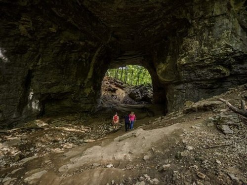 Two hikers explore a large cave entrance surrounded by rocky terrain and lush greenery.