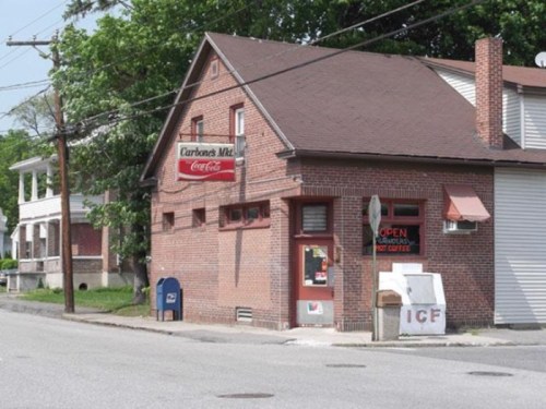 A brick building with a Coca-Cola sign, featuring a small storefront and nearby residential homes on a quiet street.