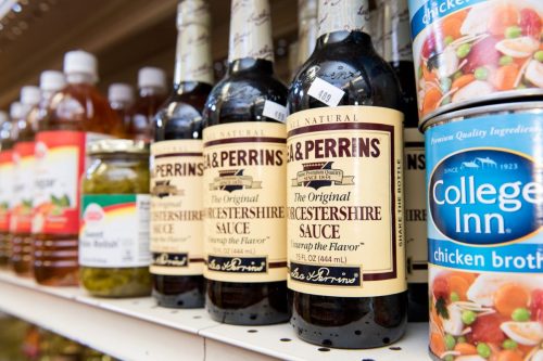 Bottles of Worcestershire sauce on a grocery store shelf, alongside jars of pickles and cans of chicken broth.