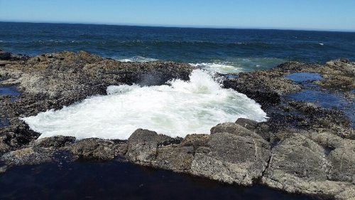Waves crashing against rocky shore with clear blue sky and ocean in the background.