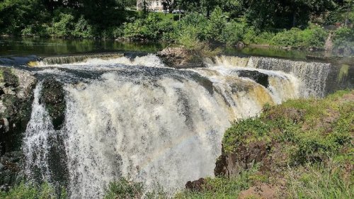 A cascading waterfall flows over rocky ledges, surrounded by lush greenery and a clear blue sky.