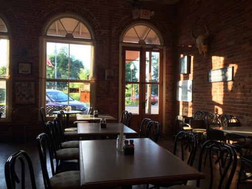 Cozy restaurant interior with wooden tables, brick walls, and large windows letting in sunlight.