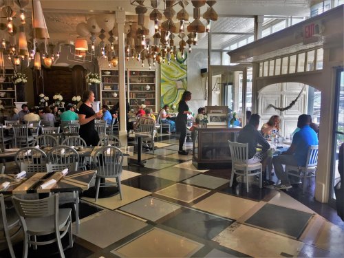 A bustling restaurant interior with diners at tables, a server, and stylish decor featuring hanging lights and plants.