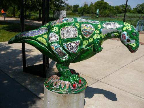 Colorful dinosaur sculpture covered in various stickers, standing on a concrete base in a park setting.