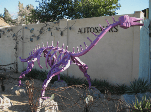 A large purple dinosaur sculpture made of metal, standing in a gravel area near a wall with "AUTOSAVERS" written on it.