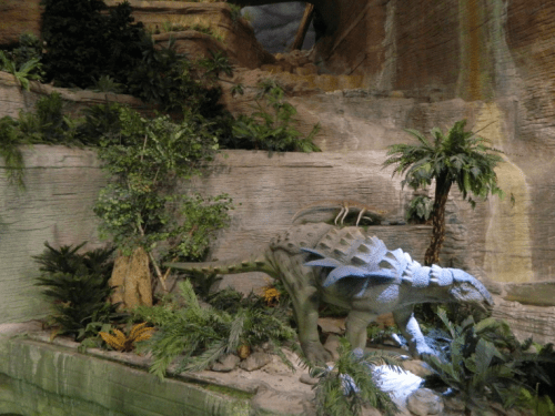 A dinosaur sculpture surrounded by lush greenery and tropical plants in a rocky environment.