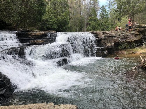 A serene waterfall cascades over rocks into a clear stream, surrounded by lush greenery and people enjoying the scenery.