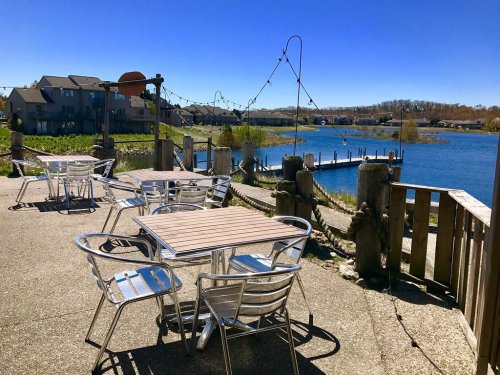Outdoor seating area by a calm blue lake, with houses in the background and clear blue skies above.