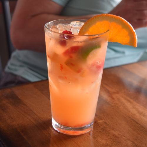 A refreshing cocktail in a tall glass, garnished with an orange slice and cherries, sitting on a wooden table.