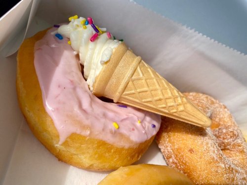 A donut topped with pink icing and a mini ice cream cone, sprinkled with colorful toppings, in a box.