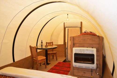 Cozy interior of a tent featuring a dining table, chairs, and a heater, with warm lighting and a rug on the floor.