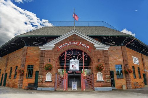 Historic Doubleday Field entrance with brick architecture, American flag, and flower planters under a blue sky.