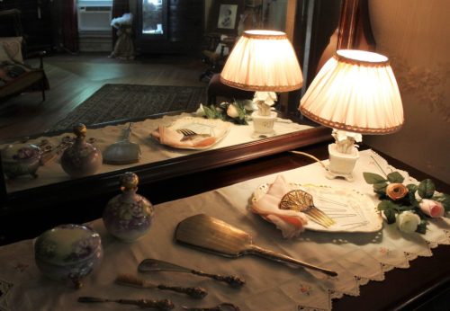 A vintage vanity with a lamp, decorative items, and grooming tools on a lace tablecloth, reflecting in a mirror.