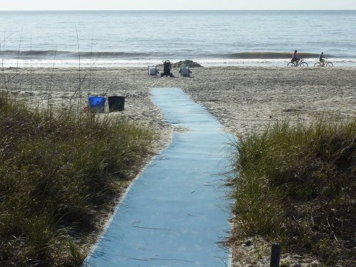 A blue pathway leads to a sandy beach with gentle waves, surrounded by grass and beachgoers on bicycles.