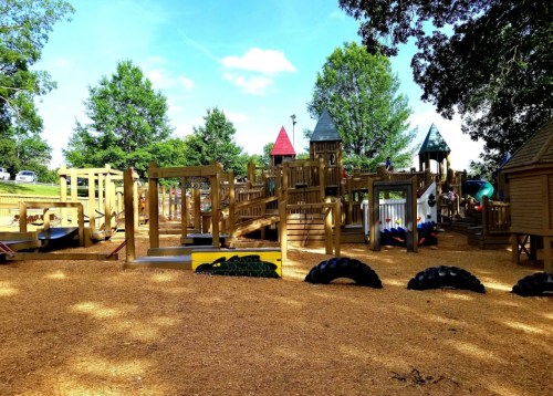 A wooden playground with slides, climbing structures, and tire swings, surrounded by trees and blue skies.
