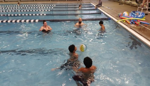 A group of people swimming and playing with a beach ball in a pool, with swim lanes visible in the background.