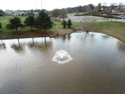 A serene pond with a fountain, surrounded by trees and a grassy area, with a person near a small dock in the background.