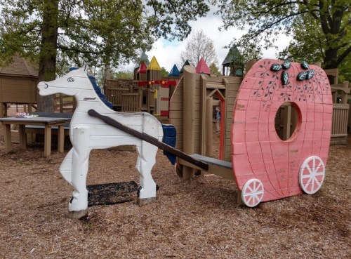 A wooden horse and cart play structure in a playground, surrounded by trees and colorful play equipment.