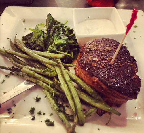 A plate featuring a seasoned steak, green beans, sautéed greens, and a small bowl of dipping sauce.
