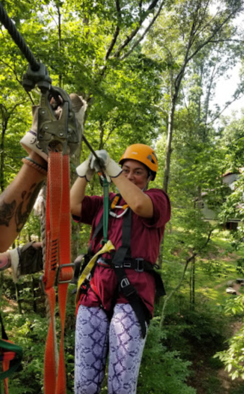 A person in a helmet and harness zip-lining through a forested area, gripping the cable with both hands.