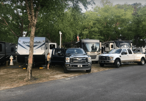 A campsite with several RVs parked, a person near a blue truck, surrounded by trees and a clear sky.