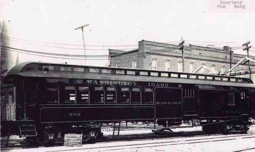 Historic black and white photo of a vintage train car labeled "Washington Idaho & Montana R.R." parked near a building.