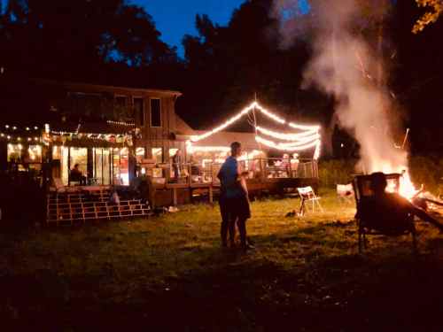 A cozy outdoor scene at night with a fire, string lights, and people gathered around a house.