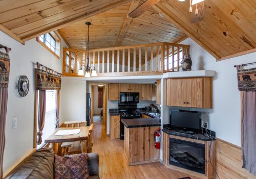 Cozy cabin interior with wooden beams, kitchen, dining area, and a loft overlooking the living space.