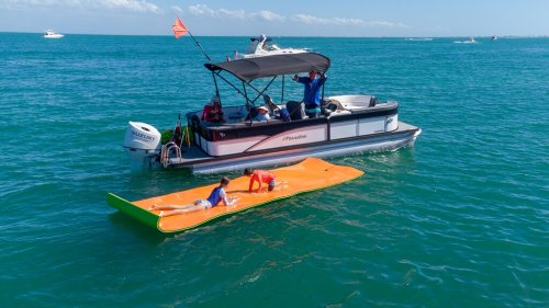 A boat with people on board near two children lounging on a large orange float in the water.