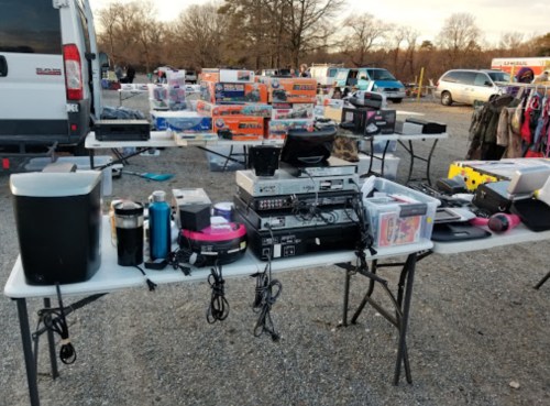 Tables filled with various items, including electronics, tools, and household goods, at an outdoor market.