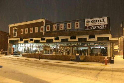 A snowy night scene of a vintage storefront with "Englanders" and "Grill" signs, featuring large windows displaying antiques.