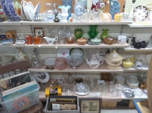 A display of various vintage glassware, ceramics, and decorative items on shelves in an antique store.
