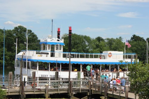 A riverboat named "Becky Thatcher" docked at a pier with people boarding, surrounded by trees and a blue sky.