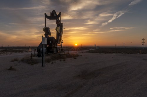 Oil pump jack silhouetted against a sunset, with a vast landscape and power lines in the background.