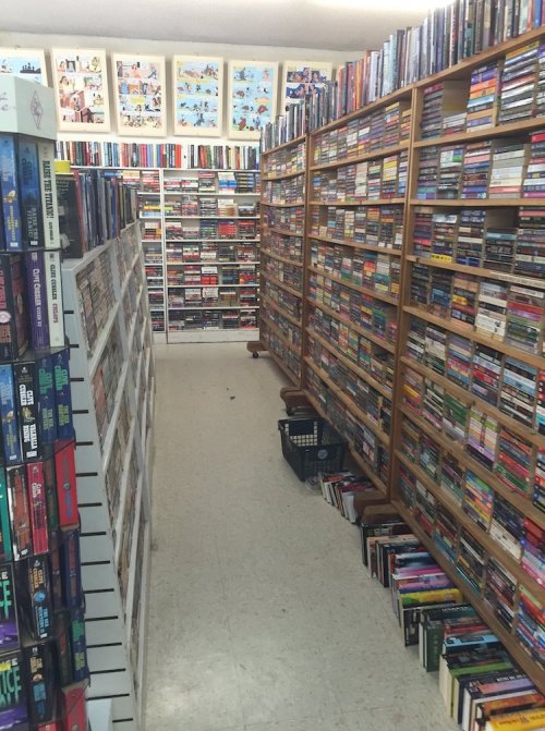 A narrow aisle between tall bookshelves filled with various books, with a few stacked on the floor and colorful artwork above.