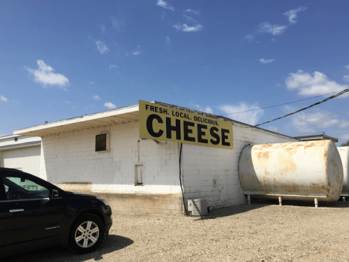 A building with a large sign reading "Fresh. Local. Delicious. Cheese." next to a parked car and a storage tank.