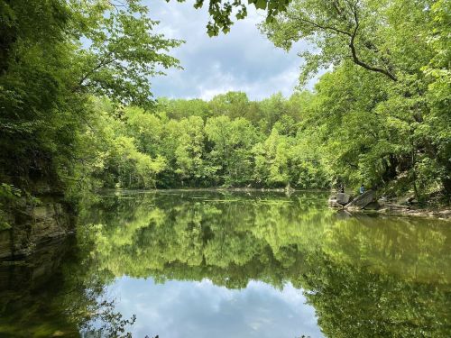 A serene lake surrounded by lush green trees, reflecting the landscape under a cloudy sky.
