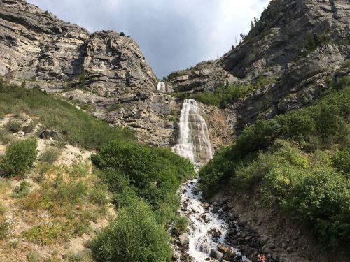 A cascading waterfall flows down rocky cliffs, surrounded by lush greenery and rugged terrain under a cloudy sky.