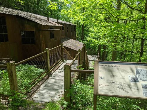 A wooden building surrounded by trees, with a pathway leading to it and an informational sign about modern technology.