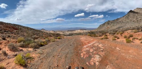 A panoramic view of a desert landscape with rocky terrain, sparse vegetation, and a cloudy blue sky.