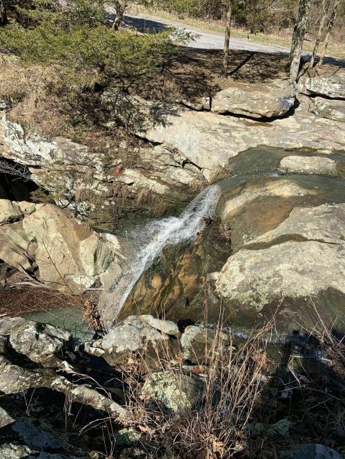 A small waterfall cascades over rocky terrain, surrounded by dry grass and trees in a natural setting.