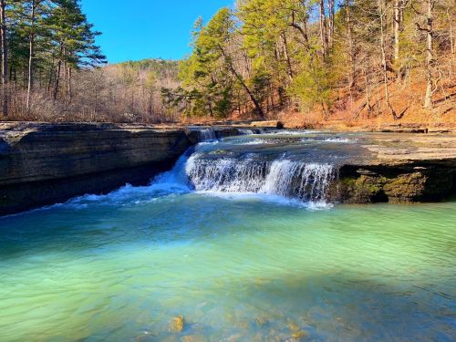 A serene waterfall cascades into a clear turquoise pool, surrounded by trees and rocky terrain under a bright blue sky.